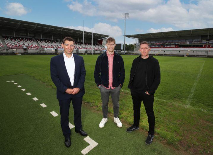 Three men stand on a rugby pitch inside an empty stadium.