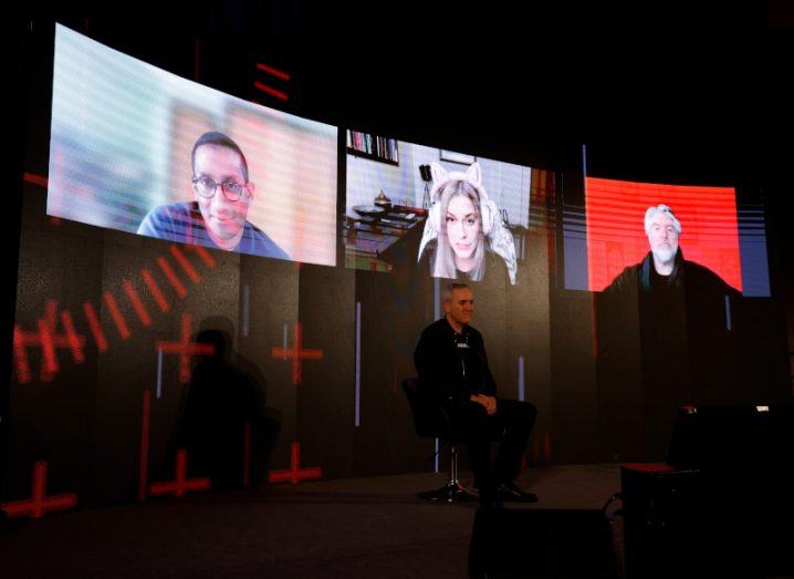 A screen with three people calling in through video call while a fourth person is seated on a stage in front of them.