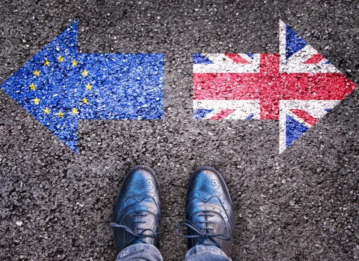 Arrow-shaped flags of the UK and the EU painted on the ground, pointing in opposite directions. A person's shoes are visible standing next to the arrows.