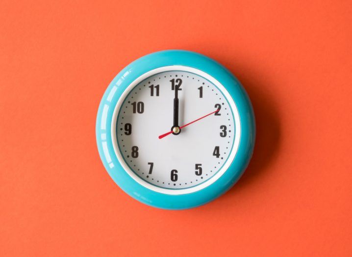 A blue clock placed on an orange wall with the time reading 12 o'clock. It signifies the 12 automation start-ups featured in this article.