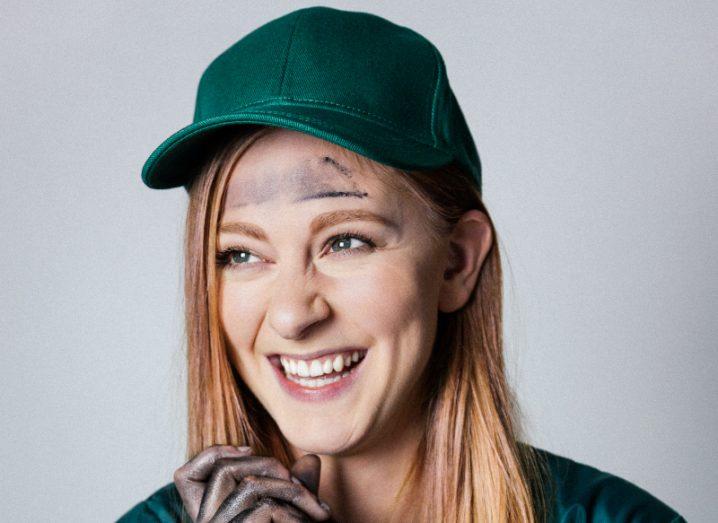 Simone Giertz in a bottle green baseball cap and coveralls, smiling. Her hands are covered in oil stains, as is her forehead.