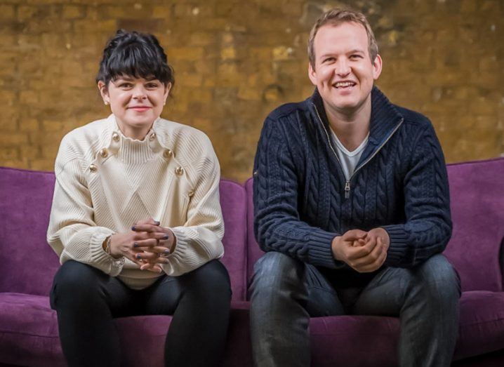 A woman and a man sitting on a purple couch with a brown wall behind them.