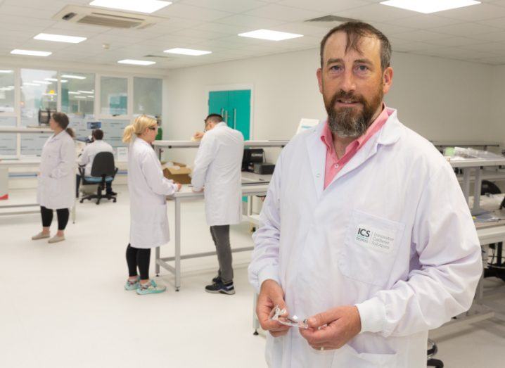 Seamus Fahey stands in a lab where people are working at benches. He is wearing an ICS Medical lab coat and holds protective eyewear in his hands.