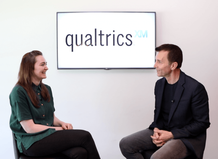 Elaine Burke and Zig Serafin sit across from each other on stools, engaged in conversation. A screen behind them displays the Qualtrics logo and XM symbol.
