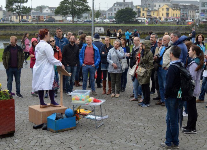 A woman in a white lab coat stands at a raised lectern on the streets of Galway next to a box of coloured balls and other props. A crowd has gathered to listen to her.