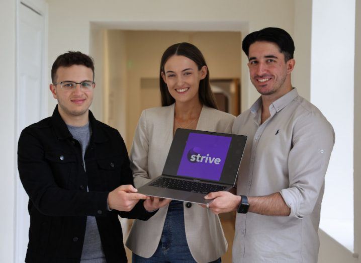 One woman and two men standing together in a white hallway, holding a laptop that has the Strive company logo on the screen.