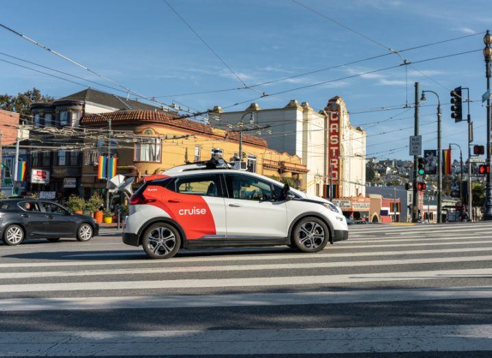 A white and red car with the Cruise logo driving in the middle of a San Francisco street with a blue sky overhead. The car is a self-driving vehicle.