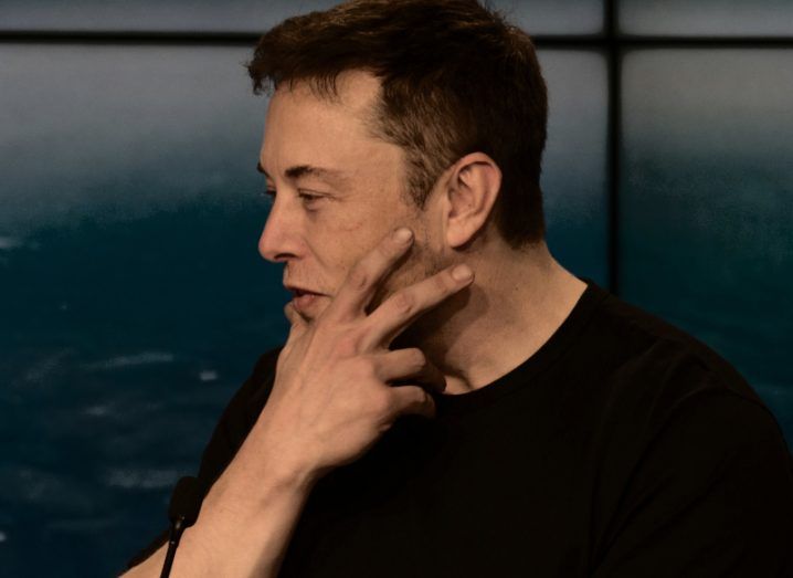A man with his hand on his chin in front of a microphone with a tiled wall behind him. He is Elon Musk.