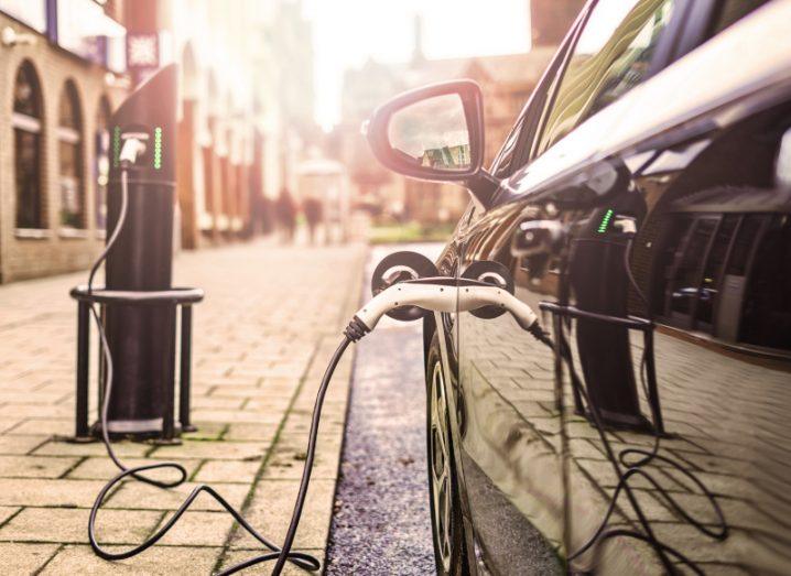 A black electric vehicle being charged next to a footpath, with a charge station visible and buildings along the side and in the background.