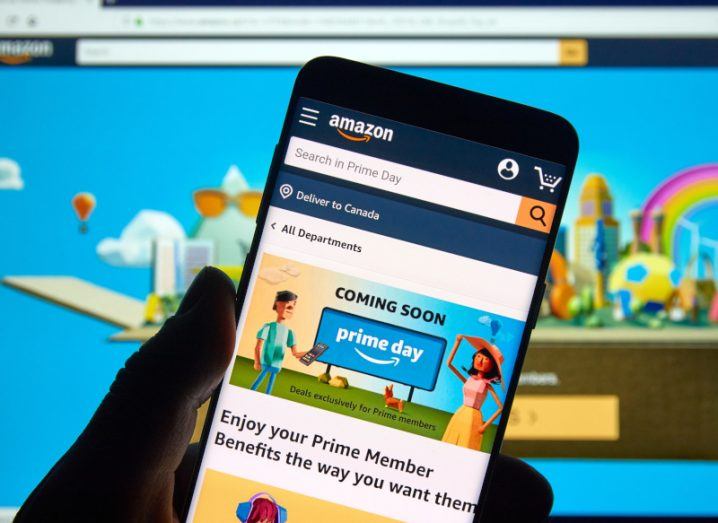 Mobile phone with an Amazon Prime Day page on the screen, with a blue Amazon screen on a computer monitor in the background.