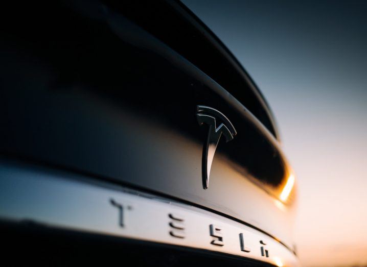 The rear of a Tesla car, showing the company's logo, against a sunset sky.