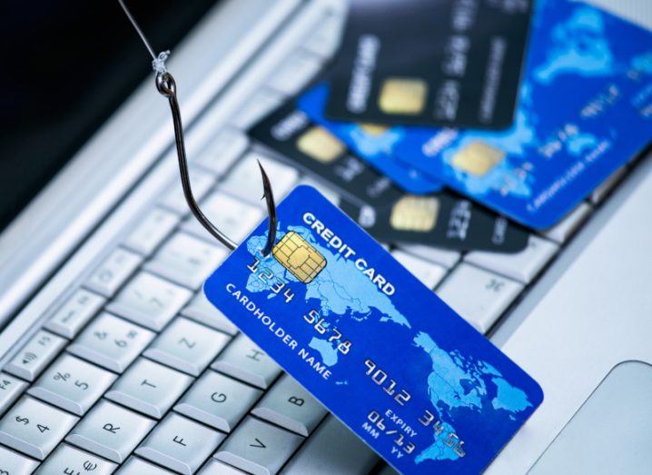 A fishing hook that is lifting up a credit card. Other credit cards are next to it and they are all resting on a laptop keyboard. The image is meant to represent phishing attacks.