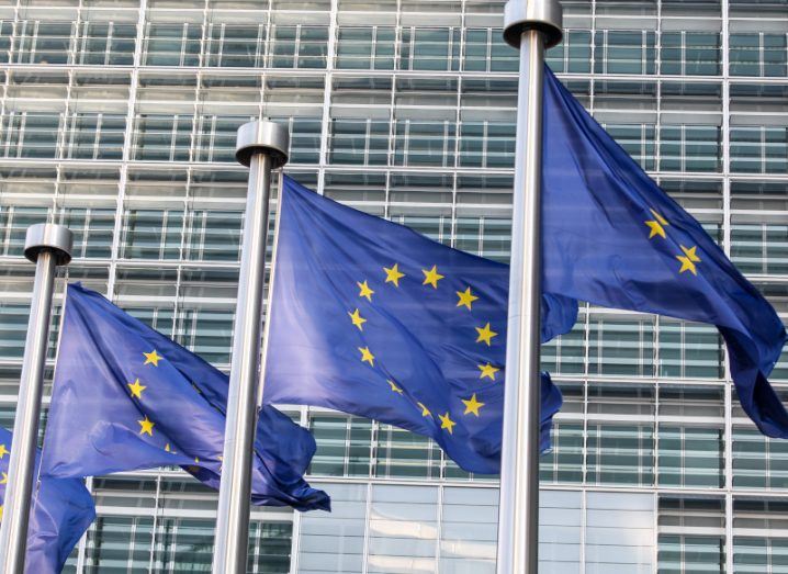 Three EU flags waving on poles outside the European Parliament building.