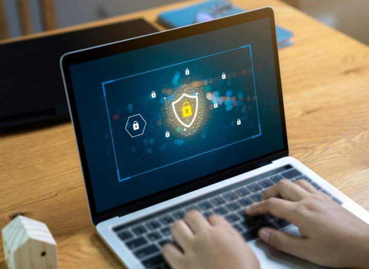 Laptop with a digital lock surrounded by a shield icon on the screen, used to represent a cybersecurity concept. The laptop is on a light brown wooden table with a small model of a house to the left.