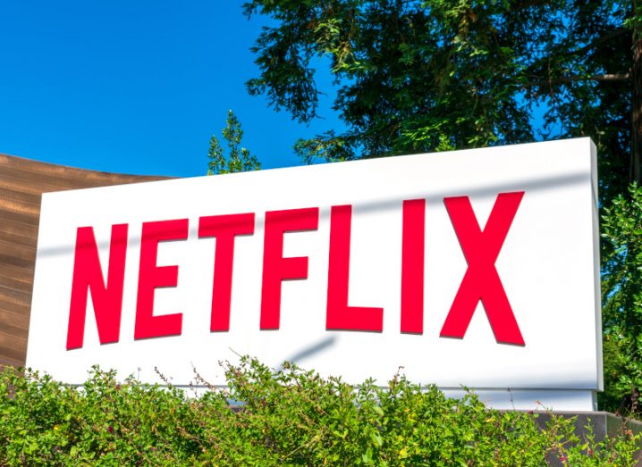 Netflix logo on a white billboard with grass in front of it and trees in the background. There is a clear blue sky overhead.