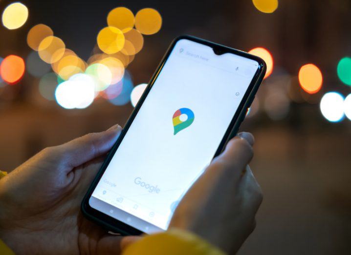 Person holding a mobile phone with the Google Maps icon in the middle of the screen. The icon is also used to show a user's current location on Maps. There are blurred city lights in the background.