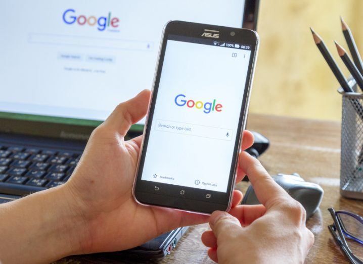 A person holding a mobile phone that has the Google logo and search function on the screen. A laptop is in the background on a desk that also has the Google logo on the screen.