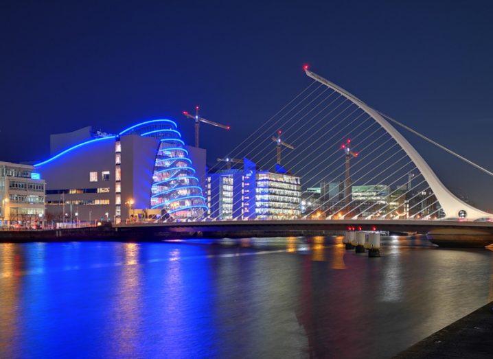 Dublin city at night with lit up buildings and a bridge over the River Liffey, with cranes visible in the distance.