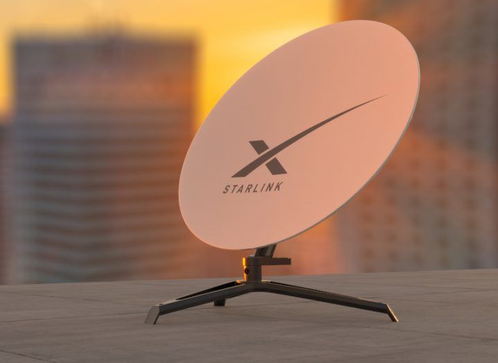 Satellite dish on a grey building roof with high-rise buildings in the background and a sunset orange sky. The dish has the Starlink name and logo on it.
