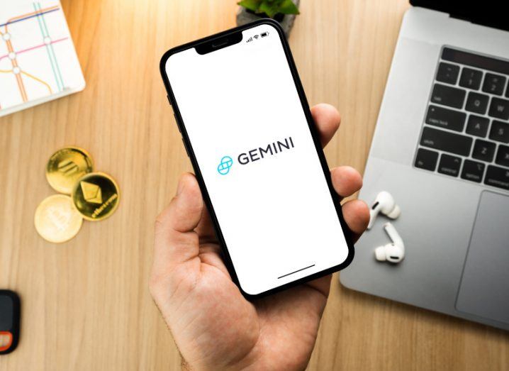 Gemini logo on mobile phone screen, being held in a person's hand above a wooden table. There is a laptop and coins used to represent cryptocurrency on the desk.