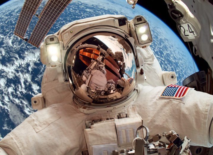 An astronaut in a space suit with a US flag from the ISS floating in space with a satellite and earth behind them.