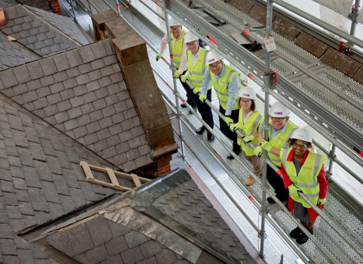 A group of people in high-vis jackets and hard hats stand on scaffolding overlooking a building project.