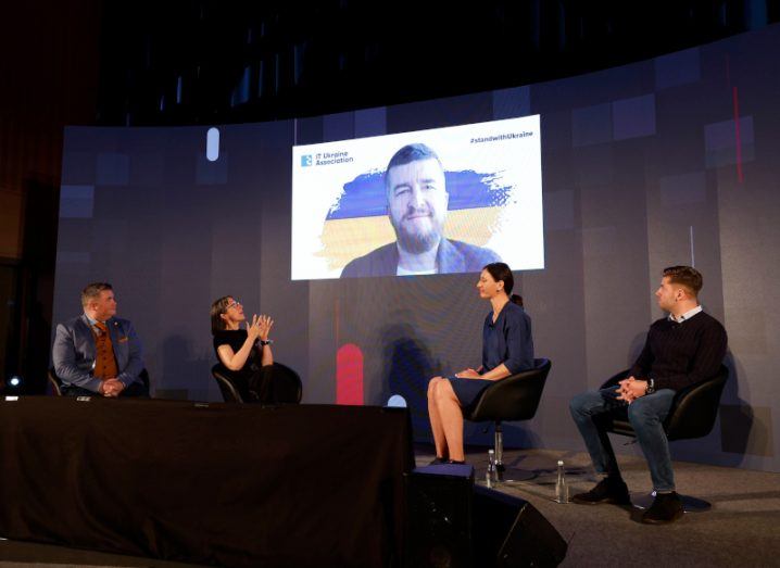 Four people sitting on chairs on a stage with a giant screen monitor showing a man speaking. They are all discussing tech in Ukraine.