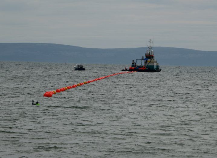 A string of orange buoys floating in a line in the sea leading to a large boat, showing a subsea cable being floated to land.