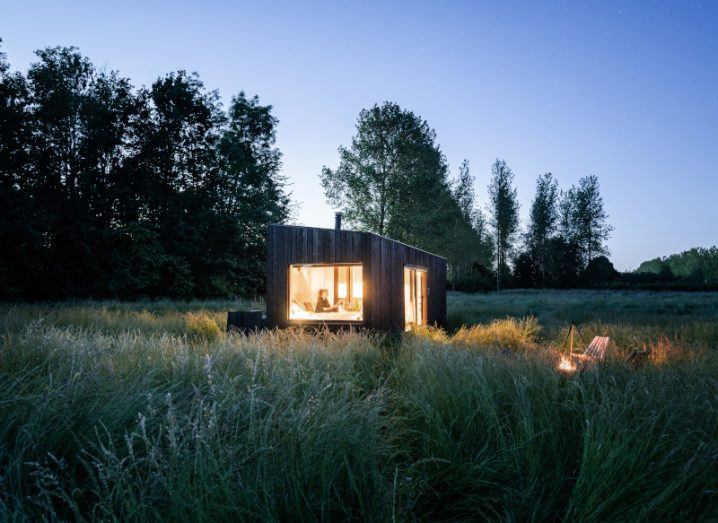 One of the Slow Cabins wooden huts set against a countryside landscape at dusk with a person inside with the light on.