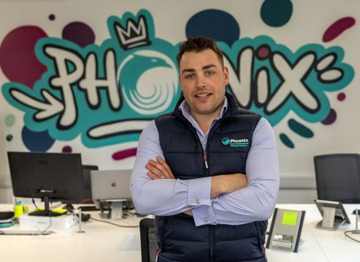 Ed Rossiter, co-CEO and founder of Phoenix, standing in an office in front of a desk with Phoenix branding on the wall behind him.
