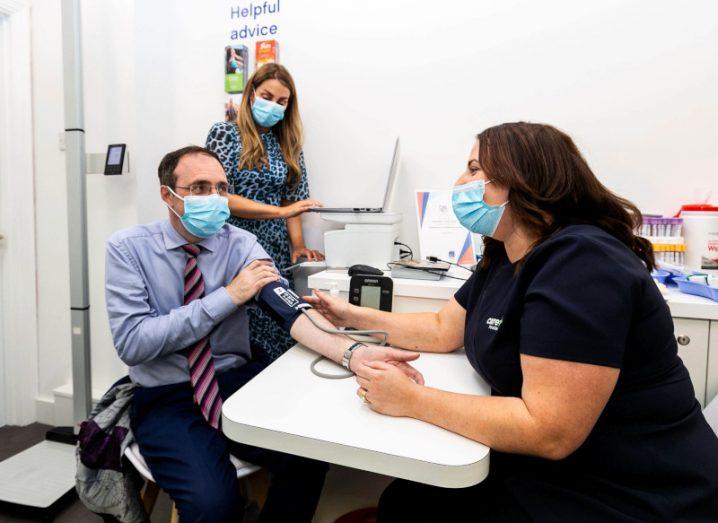 Two women and a man sitting in a pharmacy, wearing blue medical masks. The man has his left arm on the table and is getting his blood pressure taken.