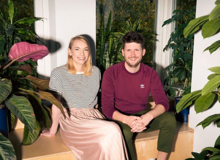 Positive Carbon founders Aisling and Mark Kirwan sit on a bench in front of a window, surrounded by large indoor plants.