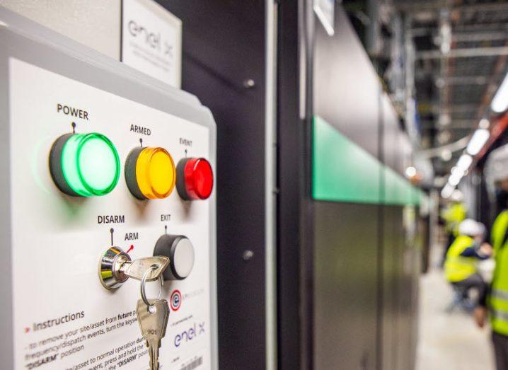 A control panel at a data centre with different coloured lights and workers visible in the background. The panel is part of Microsoft's uninterruptible power supply system.