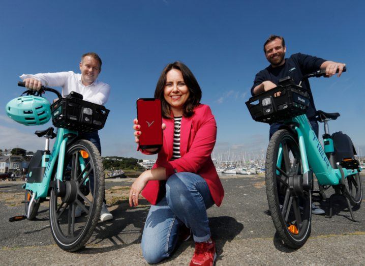 Two men and a woman with a blue sky in the background. The two men are standing next to e-bikes, while the woman is on one knee holding a phone that has the Free Now logo on it.
