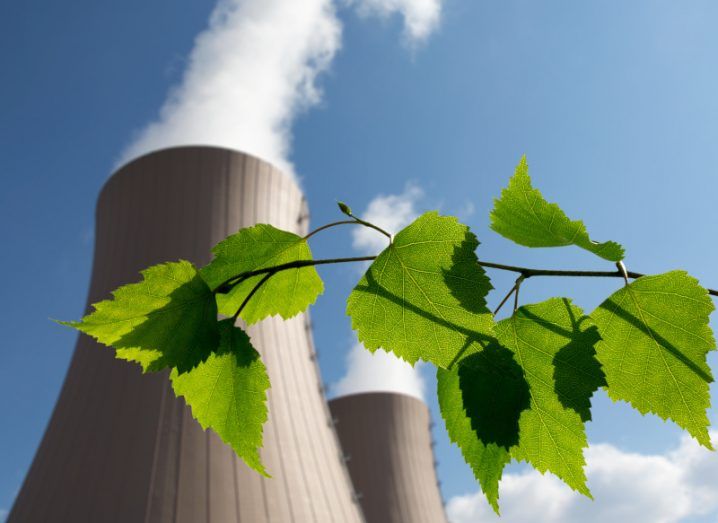 A green branch with leaves in front of a nuclear power plant, releasing smoke into a blue sky.