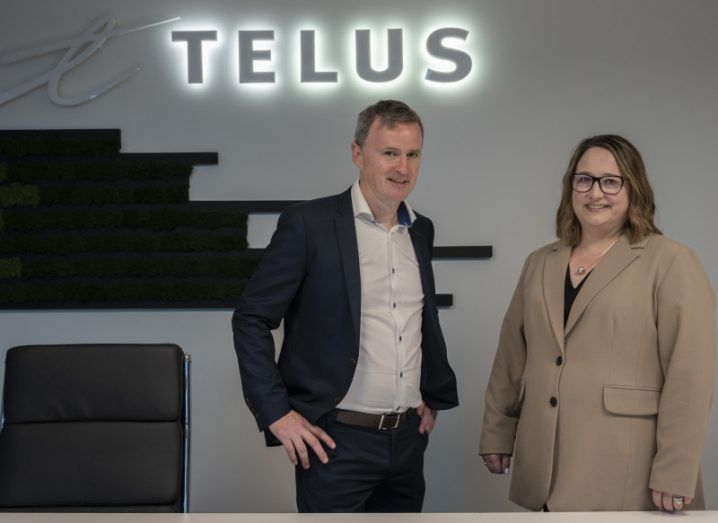 Two people stand in an office in front of a neon sign that says 'Telus'.