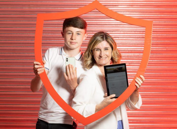 A teenage boy holding a smartphone and a woman holding a tablet posing with the Vodafone Ireland logo and branding against a red wall.