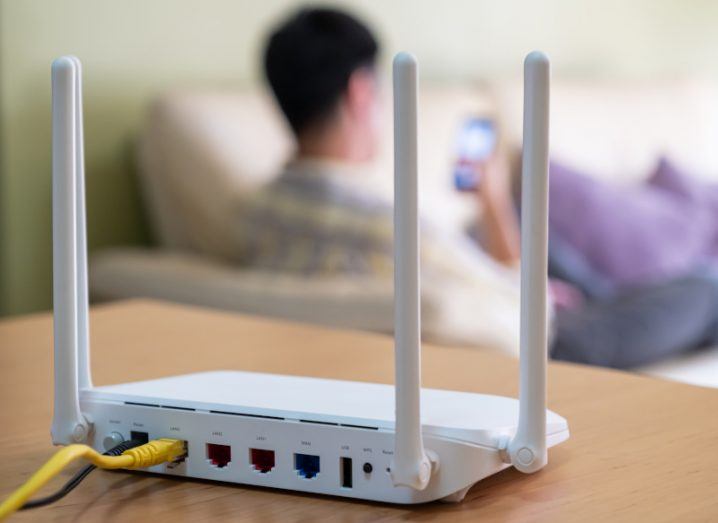 A white router with three antennae placed on a table inside a house with a person visible in the background.