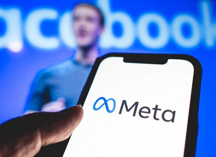 Meta logo on a smartphone screen held in a person's hand. Blurred image of Mark Zuckerberg and Facebook logo in the background.