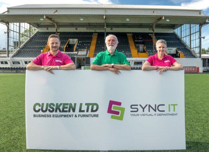 Three people standing in front of a stand in a stadium football pitch holding a sign with Sync IT and Cusken logos.