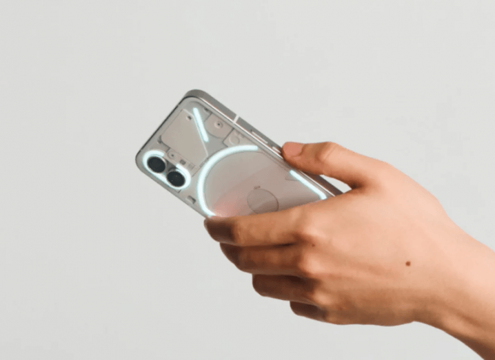 A hand holding a smartphone with lights on the back, in a grey background. It is the Nothing Phone 1.