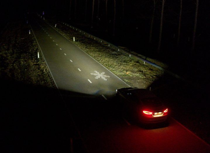A car driving on a road at night with headlights illuminating the road ahead. A snowflake symbol is being projected on the road, to warn of icy conditions ahead.