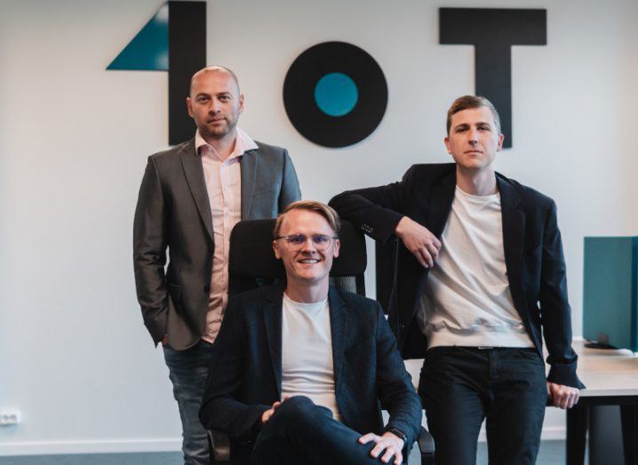 Two men standing either side of another man who is sitting on a chair, with a wall and the 1oT logo behind them.