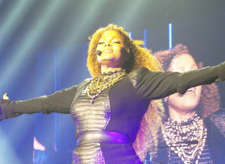 Janet Jackson on stage with her arms out to her side, with a microphone next to her head. A screen with Janet Jackson is visible in the background.