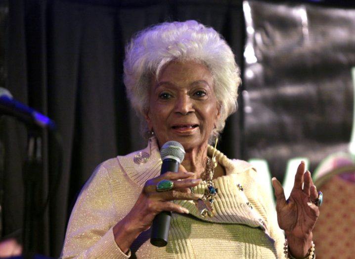 A woman holding a microphone in her hand speaking on a stage. She is Nichelle Nichols.