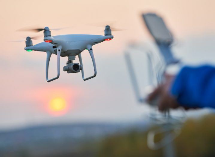 A drone in flight against a setting sun. The hands of the drone's remote operator are visible in the foreground.