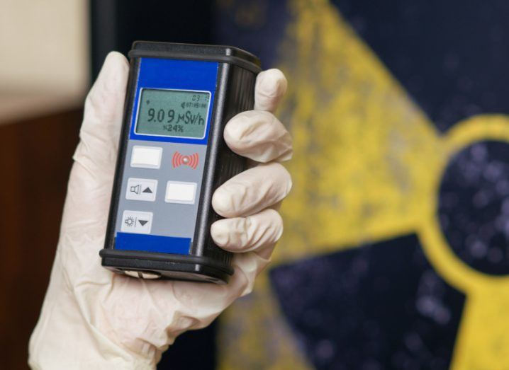 A gloved hand holding a geiger counter with a radioactive symbol in the background.