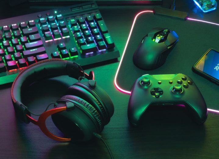 A pair of headphones, a mouse and a gaming console laying on a table, with a keyboard next to them.