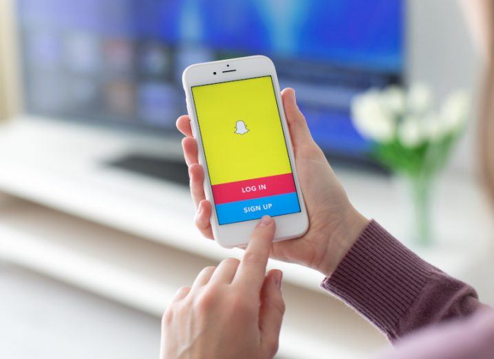 Person holding a smartphone with the Snapchat logo on the screen, in a living room. The options to log in or sign up are visible on the phone.