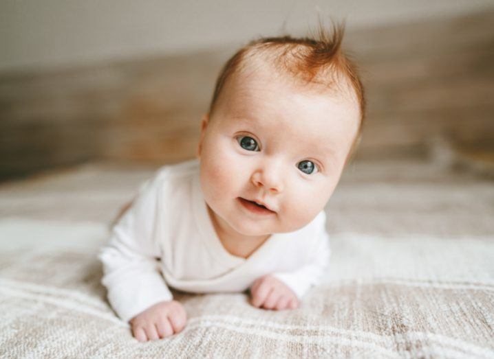 A newborn baby crawls on a woven blanket. Their eyes are wide and red hair slightly tousled.